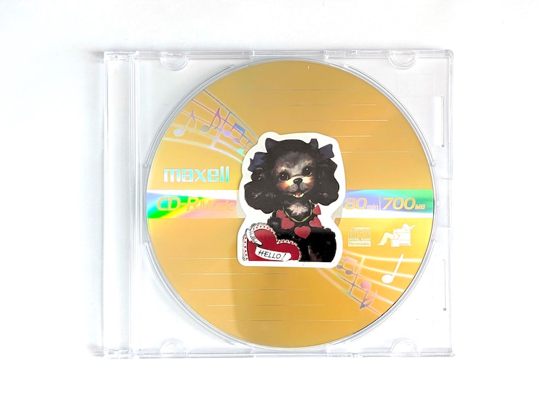 May include: A Maxell CD-R disc in a clear plastic case. The gold disc has a sticker of a black dog with a red heart collar and a heart-shaped sign that says "Hello!". The disc has musical notes printed on it.