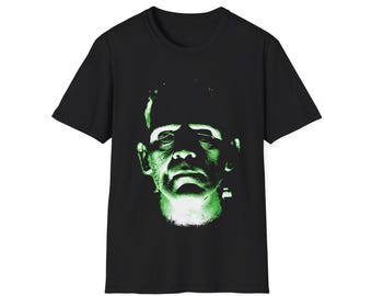 May include: Black t-shirt featuring a green and white graphic of Frankenstein's monster. The design is a close-up of the monster's face, with a focus on the eyes and forehead. The t-shirt is made of a soft material and has a classic crew neck.