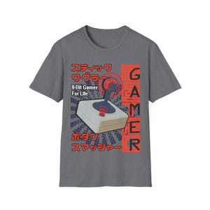 Puede incluir: Una camiseta gris con un gráfico de mando de videojuego retro. El diseño incluye la palabra "Gamer" en rojo, texto japonés y la frase "8-Bit Gamer For Life."