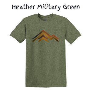 Puede incluir: Camiseta verde militar jaspeado con un gr&aacute;fico de monta&ntilde;a en tonos marrones y naranjas. La camiseta es de estilo cuello redondo y manga corta. El texto "Heather Military Green" est&aacute; encima de la camiseta.