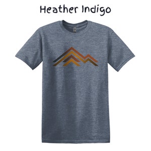 Puede incluir: Camiseta Heather Indigo con un gr&aacute;fico de monta&ntilde;a. La camiseta de manga corta presenta un dise&ntilde;o de cordillera en tonos marrones, naranjas y grises. El texto "Heather Indigo" est&aacute; encima de la camiseta en una fuente manuscrita.
