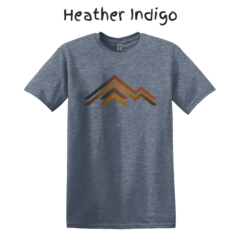 Puede incluir: Camiseta Heather Indigo con un gr&aacute;fico de monta&ntilde;a. La camiseta de manga corta presenta un dise&ntilde;o de cordillera en tonos marrones, naranjas y grises. El texto "Heather Indigo" est&aacute; encima de la camiseta en una fuente manuscrita.