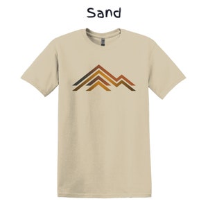 Puede incluir: Camiseta color arena con un dise&ntilde;o gr&aacute;fico de monta&ntilde;a. El dise&ntilde;o de la monta&ntilde;a presenta l&iacute;neas horizontales marrones, naranjas y grises. La palabra "Sand" est&aacute; impresa encima de la camiseta.
