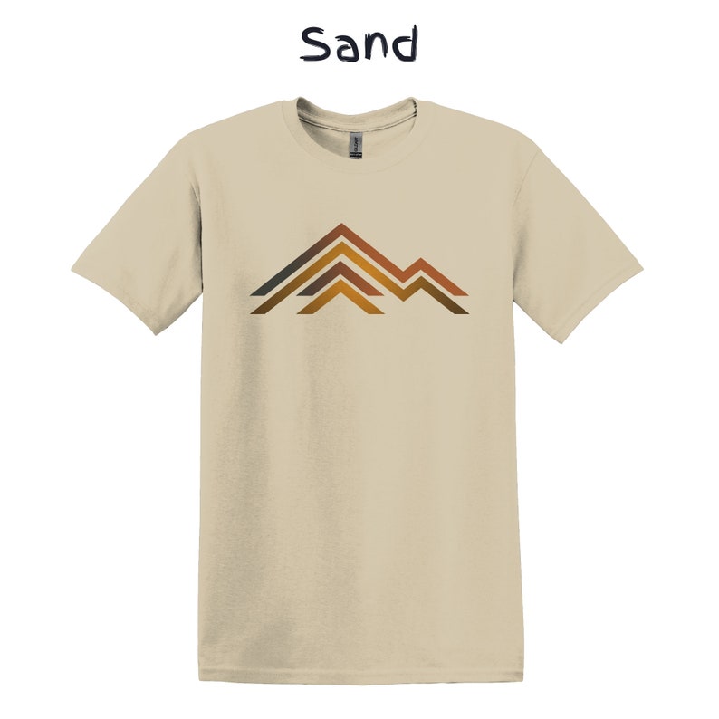 Puede incluir: Camiseta color arena con un dise&ntilde;o gr&aacute;fico de monta&ntilde;a. El dise&ntilde;o de la monta&ntilde;a presenta l&iacute;neas horizontales marrones, naranjas y grises. La palabra "Sand" est&aacute; impresa encima de la camiseta.