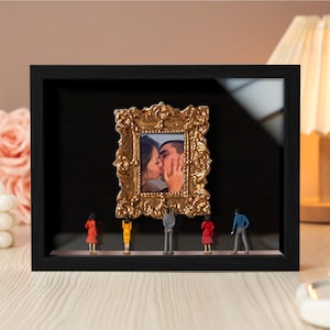 May include: A black shadow box frame with a gold ornate picture frame inside, displaying a photo of a couple kissing. Miniature figures in various outfits stand below the frame. The background is black.