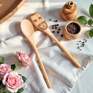 May include: Two wooden spoons, one engraved with a smiling face, rest on a white cloth. Pink roses and a small wooden bowl with peppercorns are also present. A wooden container with a lid and a wooden bowl with peppercorns are also present.