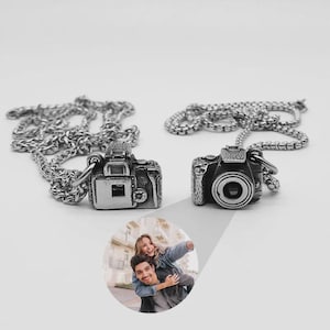 May include: Two silver-colored camera pendant necklaces. Each pendant is a detailed miniature camera attached to a chain. The necklaces are displayed on a white surface. The image also includes a photo of a couple.