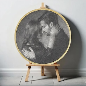 May include: A circular string art portrait in black and white, depicting a couple in an embrace, within a gold-coloured frame. The artwork is displayed on a wooden easel against a white wall.