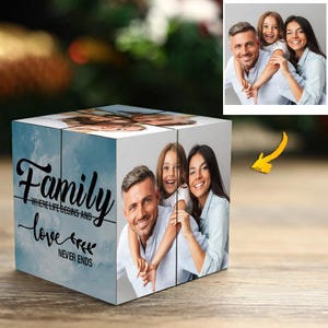 Op de afbeelding: Een gepersonaliseerde fotokubus met familiefoto's en de woorden "Family, where life begins and love never ends" in zwart schrift tegen een blauwe hemelachtergrond. De kubus toont meerdere familiefoto's.
