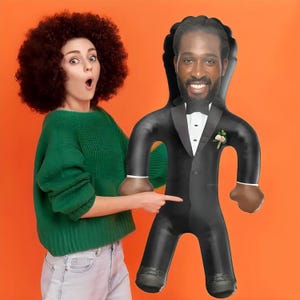 May include: An inflatable groom doll wearing a black tuxedo with a white shirt, bow tie, and boutonniere. The doll has a smiling face and is held by a person wearing a green sweater and light blue jeans against an orange background.
