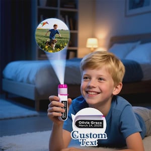 May include: A young boy smiles while holding a white and pink projector flashlight. The flashlight projects an image of a boy, dog, and kite. The flashlight has the text "Olivia Grace March 14, 2025" and "Custom Text".