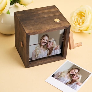 May include: A dark brown wooden photo cube with a gold-colored button on top and a photo of two women. A small wooden stand and a photo are next to the cube. Yellow flowers are in the background.