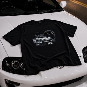May include: A black short-sleeve t-shirt featuring a graphic print of a white sports car drifting with smoke, a tachometer gauge, the number 94 in a square box, and the Japanese characters 夜走 printed in white on the front center.