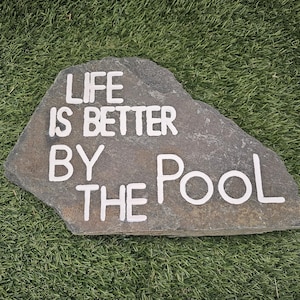 May include: A gray stone with the phrase "LIFE IS BETTER BY THE POOL" in white lettering. The stone is set against a backdrop of green artificial turf, suggesting an outdoor or poolside setting.