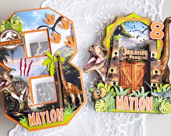 May include: Two dinosaur-themed birthday cake toppers. One is a number 8 with a green dinosaur and a Jurassic Park sign. The other is a number 8 with a dinosaur and a palm tree. Both toppers have the name Waylon on them.