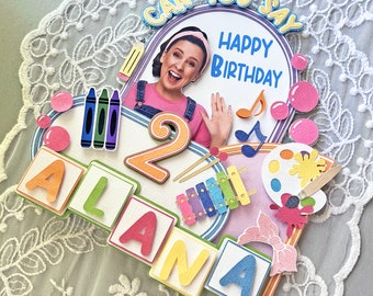 May include: A colourful birthday cake topper featuring a girl's face, the number 2, the name Alana, and the words "Can You Say Happy Birthday".