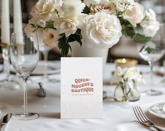 Puede incluir: Una maqueta de tarjeta blanca con el texto "QUICK MOCKUPS BOUTIQUE" en una mesa. La mesa está decorada con un arreglo floral, velas y cristalería. La tarjeta es para la colocación del diseño.