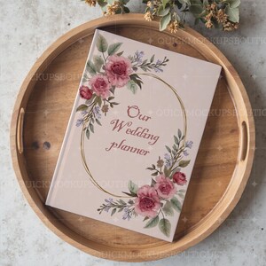 May include: A pink wedding planner with a gold floral wreath and the text "Our Wedding Planner" on the cover. The planner is sitting on a wooden tray.