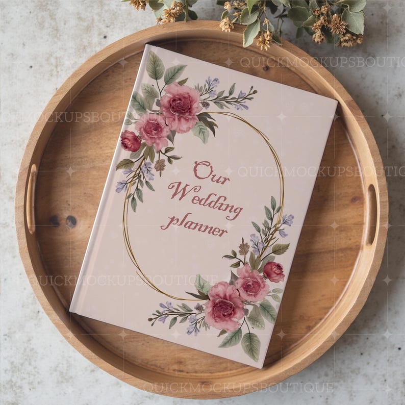 May include: A pink wedding planner with a gold floral wreath and the text "Our Wedding Planner" on the cover. The planner is sitting on a wooden tray.