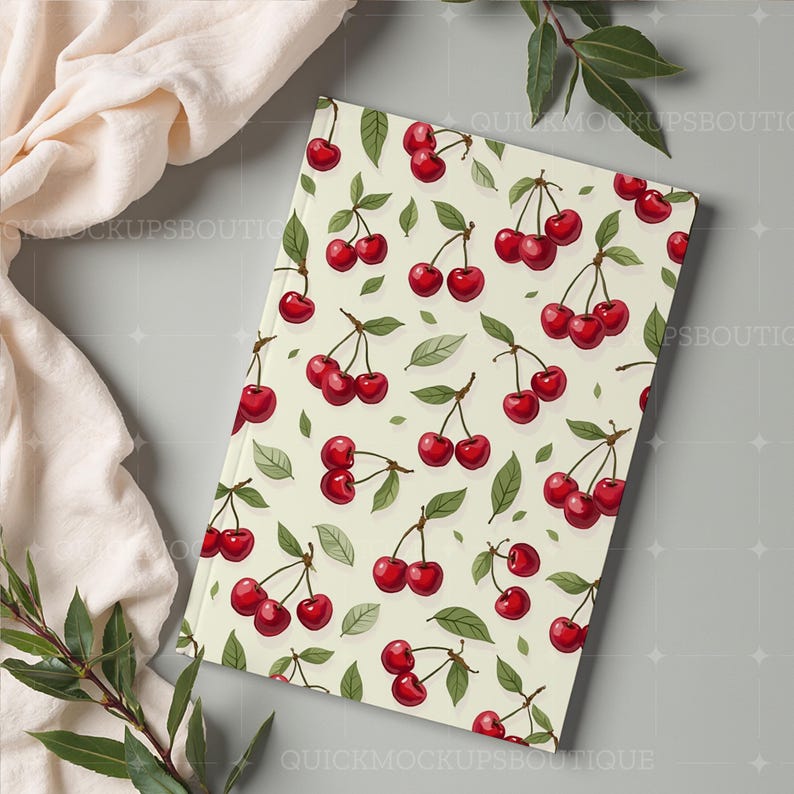 May include: A cream-colored notebook with a red cherry pattern. The notebook is on a gray surface with a white cloth and green leaves.