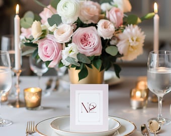 Puede incluir: Un centro de mesa floral rosa y blanco con un jarrón dorado se encuentra sobre un mantel blanco. Una cubertería con un plato blanco, borde dorado y cubiertos está sobre la mesa. Una pequeña tarjeta con el número "No. 2" impreso en ella está delante del plato.