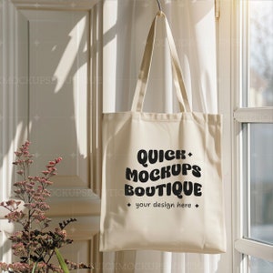 Op de afbeelding: Een witte canvas draagtas met zwarte tekst die "Quick Mockups Boutique" en "your design here" zegt. De tas hangt aan een haak voor een raam met witte gordijnen.