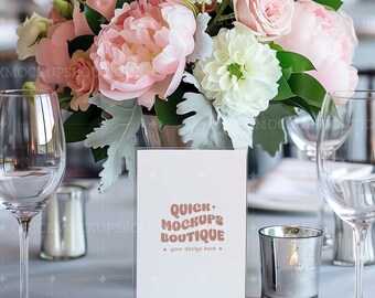 Puede incluir: Una tarjeta blanca con el texto "Quick Mockups Boutique" en una fuente retro marrón. La tarjeta está en un marco de acrílico transparente y está sentada en una mesa con un ramo de flores rosas y blancas, dos copas de vino y un portavelas plateado.