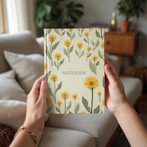 May include: A white notebook with a floral pattern of yellow flowers and green leaves. The notebook has the word "NOTEBOOK" printed in black on the front cover.