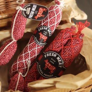 Puede incluir: Una cesta de mimbre llena de varias salchichas de ganchillo rojas y blancas. Cada salchicha está envuelta en una red blanca y tiene una etiqueta negra que dice "FRESH MEAT" con una ilustración de vaca. La cesta contiene papel marrón.