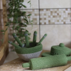 May include: A collection of green and gray crocheted kitchen toys, including a rolling pin, bowl with utensils, and a covered dish. The toys are arranged on a wooden cutting board, with a sprinkle of white powder. The background features a tiled wall and greenery.