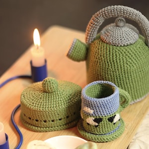 May include: A handmade crochet tea set in green and grey, comprising a teapot, cup, and lid, displayed on a wooden tray. A small white dish holds two biscuits. A lit candle in a blue holder provides a warm light.
