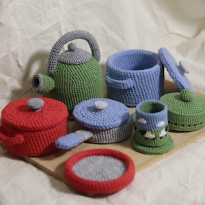 Può includere: Una collezione di articoli da cucina giocattolo all'uncinetto, tra cui un bollitore verde, pentole rosse e blu con coperchi, una padella e una tazza con un disegno di pecora. Gli oggetti sono disposti su un vassoio di legno.
