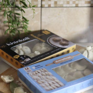 May include: Two boxes of dumplings, one labeled "khinkali" and the other "dumplings with farm meat", sit on a wooden cutting board. The "khinkali" box is black and yellow, while the other is blue. The boxes contain multiple dumplings.