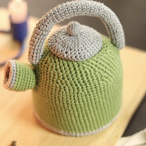 May include: A handmade, green crochet teapot with a gray handle, lid, and trim. The teapot is sitting on a wooden surface. A lit candle is in the background.