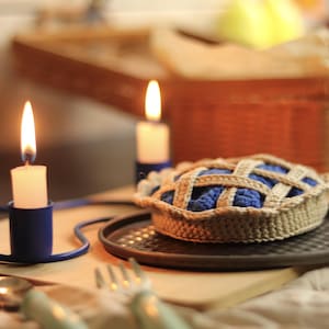 May include: A crocheted pie with a lattice crust, filled with blue yarn, sits on a dark plate. Two lit candles in blue holders and silverware are in the foreground. A wooden cutting board and a wicker basket are in the background.