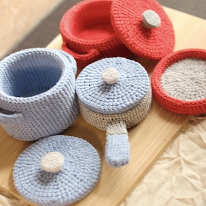 Op de afbeelding: Een verzameling handgemaakte gehaakte speelgoed potten en pannen in verschillende kleuren. De set bevat een rode pot met deksel, een lichtblauwe pot met handvatten en een blauw-grijze pan met een handvat. De deksels hebben kleine, crèmekleurige knoppen.
