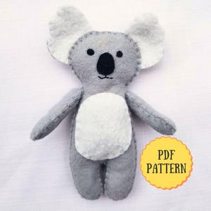 Op de afbeelding: Een handgemaakt grijze vilten koala beer knuffel met witte accenten op de oren en buik. De koala heeft een zwarte neus en ogen. De tekst "PDF PATTERN" staat in een gele cirkel.
