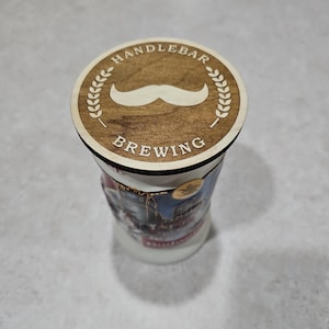 Könnte beinhalten: Ein Untersetzer aus Holz mit braunem Hintergrund und einem weißen Schnurrbart mit einem Lorbeerkranz-Design. Der Text "HANDLEBAR BREWING" ist auf den Untersetzer gedruckt.