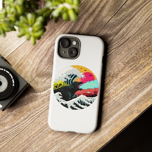 May include: A white iPhone case with a colorful graphic of Godzilla breathing fire in front of a large wave. The graphic is in a circle with a yellow, blue, and red color scheme.