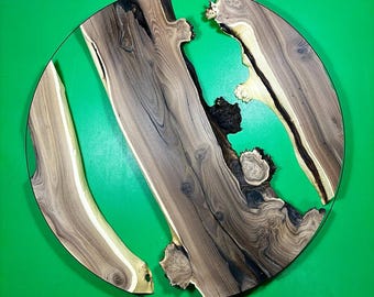 May include: A round wooden table top with a green epoxy resin inlay. The wood has a natural brown color with dark brown grain patterns. The epoxy resin is a bright green color and flows through the wood in a natural, organic shape.