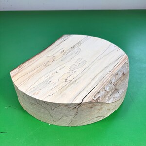 Puede incluir: Un bloque de madera semicircular de color claro, con un aspecto natural sin terminar. La veta de la madera es visible, con una mezcla de tonos claros y oscuros. El bloque tiene una base plana y una parte superior curva, con un borde rugoso similar a la corteza.
