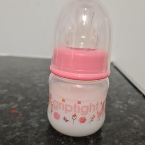 Peut inclure: Un biberon rose pour b&eacute;b&eacute; avec un couvercle transparent et une &eacute;tiquette blanche sur laquelle est inscrit "griptight" avec un motif de papillon et de fleurs.