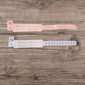 May include: Two blank hospital identification bracelets, one pink and one light blue, with spaces for name, bed number, sex, and date. The bracelets are on a wooden surface.
