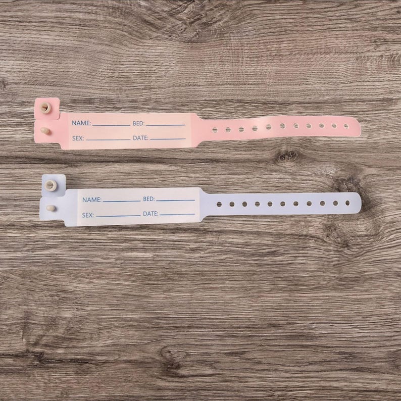 May include: Two blank hospital identification bracelets, one pink and one light blue, with spaces for name, bed number, sex, and date. The bracelets are on a wooden surface.