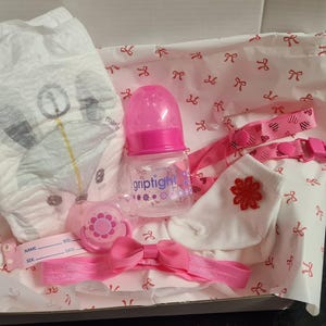 May include: A pink and white baby gift set with a diaper, a bottle, a pacifier, a headband, and a bib. The items are arranged in a white box with a pink and white patterned lining.