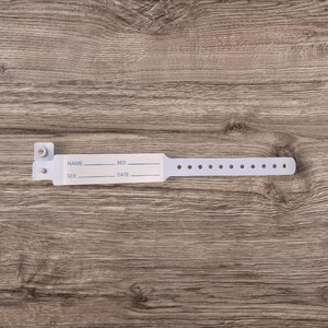 May include: White plastic hospital identification bracelet with fields for name, bed number, sex, and date. The bracelet has a series of small holes along the edge.