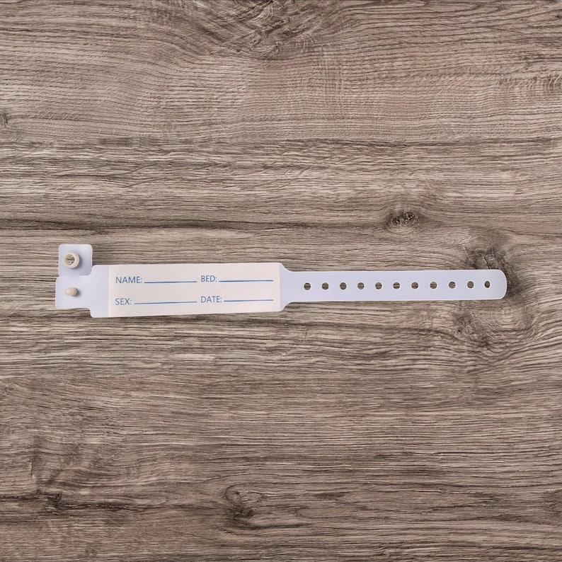 May include: White plastic hospital identification bracelet with fields for name, bed number, sex, and date. The bracelet has a series of small holes along the edge.