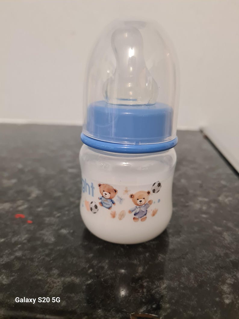 Peut inclure: Un biberon transparent avec un couvercle bleu et un motif bleu et blanc repr&eacute;sentant deux ours en peluche jouant au football. Le biberon a une t&eacute;tine en silicone transparente.