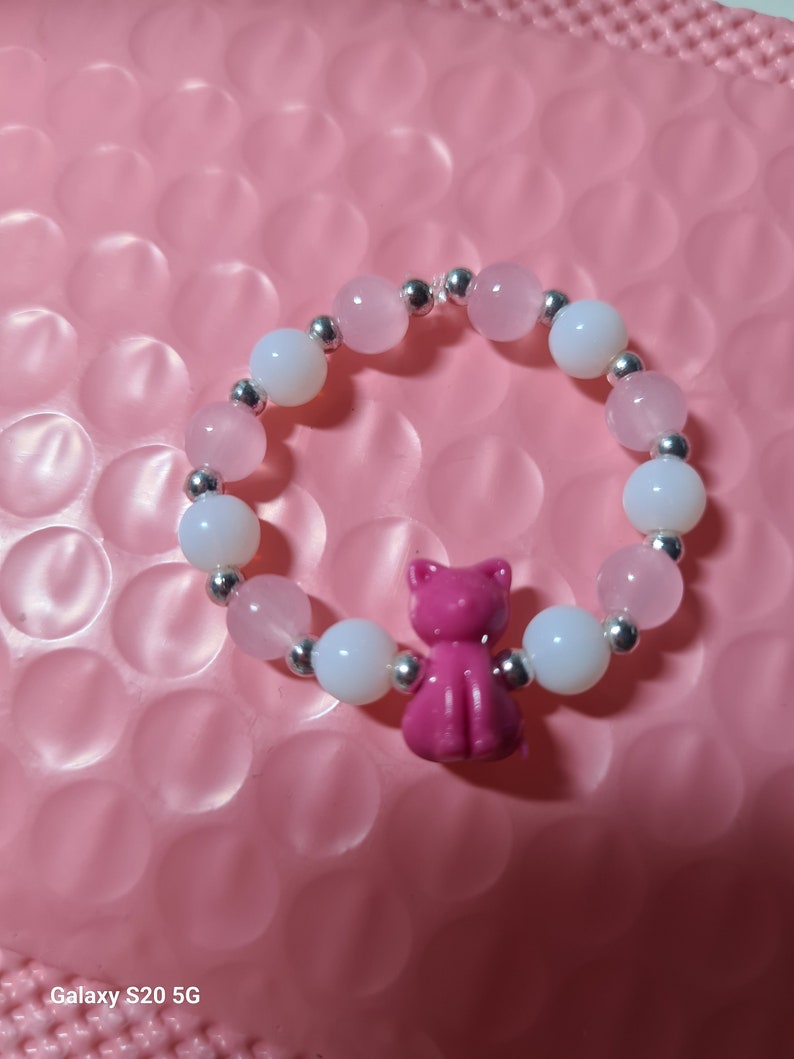 K&ouml;nnte beinhalten: Ein rosa-wei&szlig;es Perlenarmband mit einem rosa Katzenanh&auml;nger. Das Armband besteht aus kleinen runden Perlen und silbernen Zwischenst&uuml;cken.