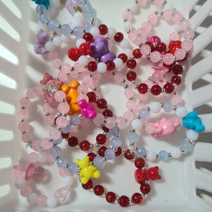 K&ouml;nnte beinhalten: Eine Sammlung bunter Perlenarmb&auml;nder mit verschiedenen Anh&auml;ngern, darunter B&auml;ren, Herzen und Sterne. Die Armb&auml;nder sind aus rosa, roten, blauen und wei&szlig;en Perlen gefertigt.